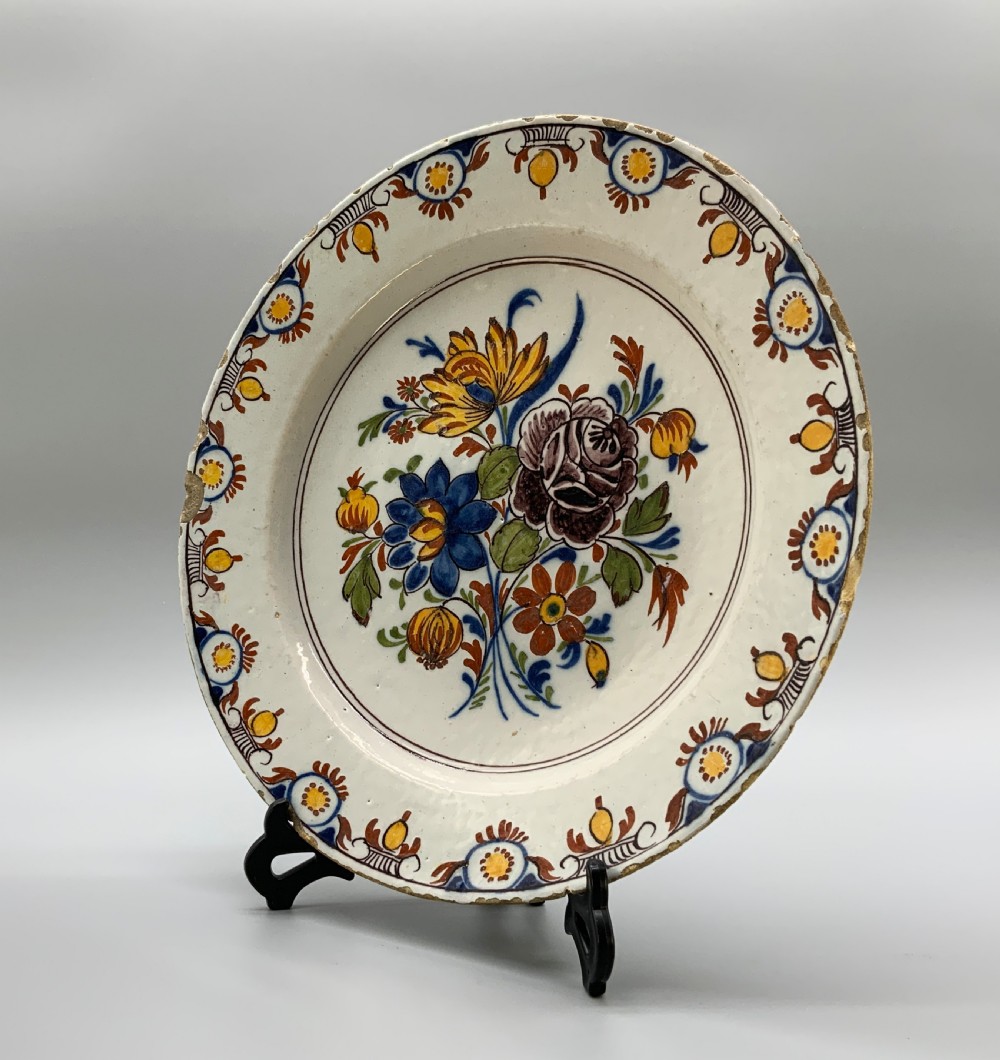 An 18th-century circular tin-glazed earthenware charger plate decorated with hand-painted floral motifs in manganese purple, cobalt blue, ochre yellow, and green. View XI