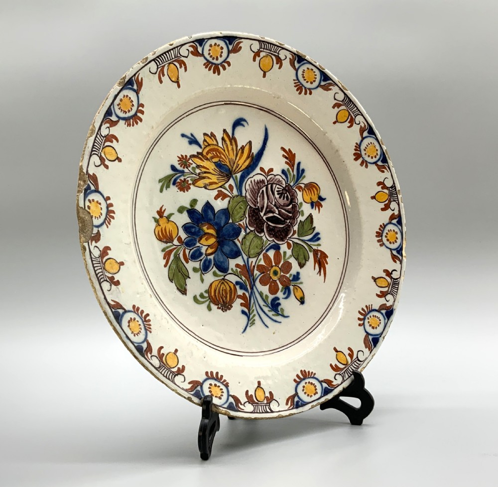 An 18th-century circular tin-glazed earthenware charger plate decorated with hand-painted floral motifs in manganese purple, cobalt blue, ochre yellow, and green. View XII