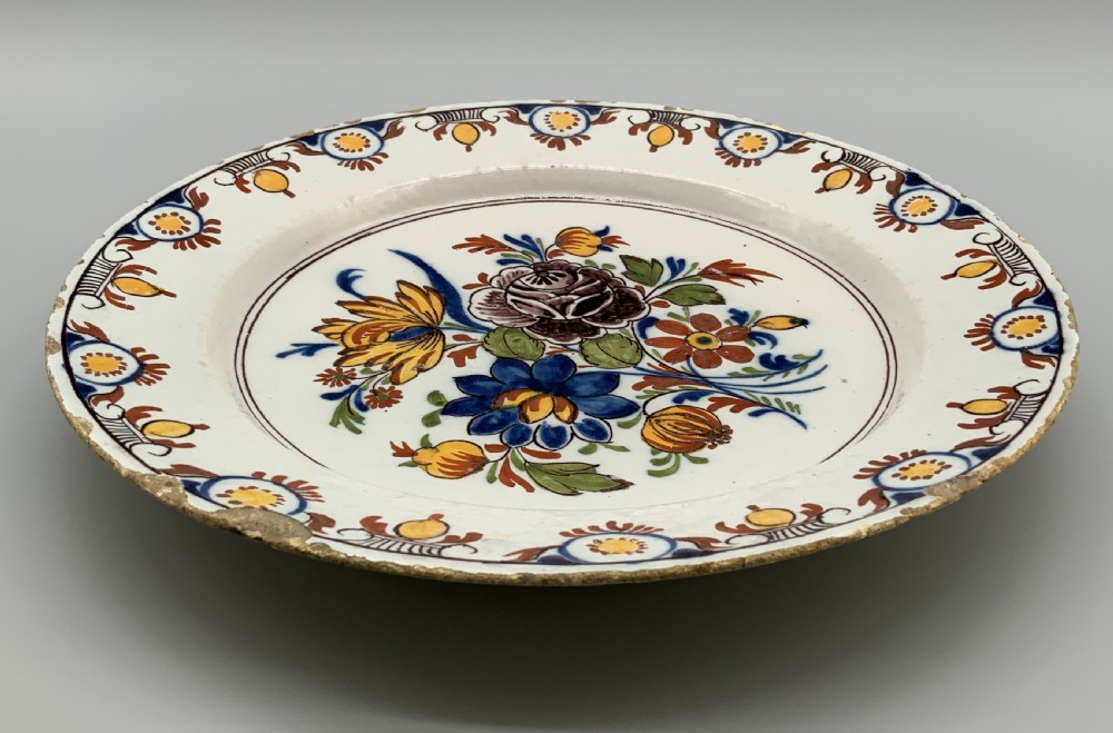 An 18th-century circular tin-glazed earthenware charger plate decorated with hand-painted floral motifs in manganese purple, cobalt blue, ochre yellow, and green. View XV