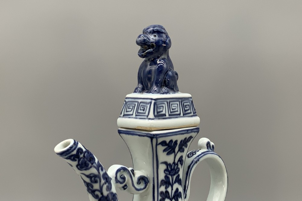 A blue and white porcelain ewer decorated with hand-painted floral foliage, featuring a high curved handle, slender curved spout, and a cover with a Foo dog finial. View X