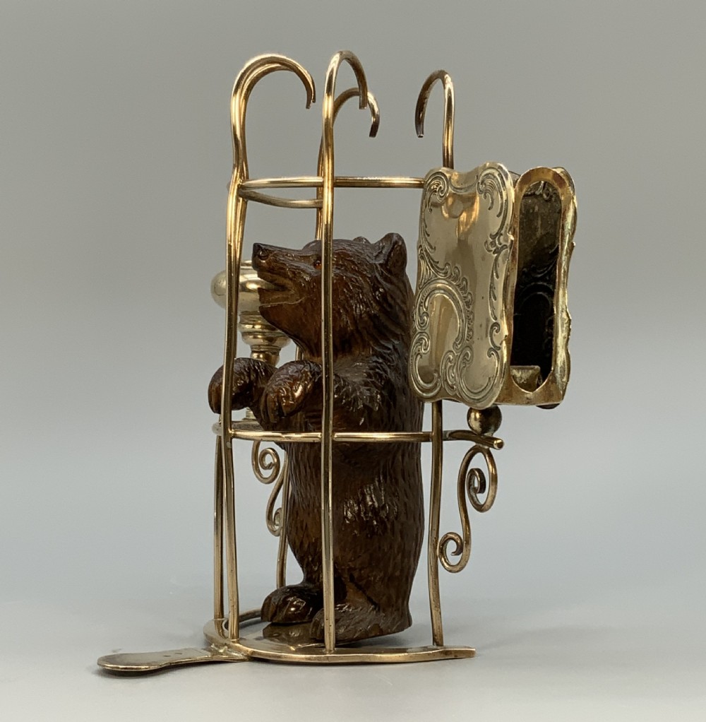 A 19th-century carved wooden bear standing upright inside a brass wire cage, attached to a brass candlestick holder on the left and an engraved brass vesta case on the right. View IV