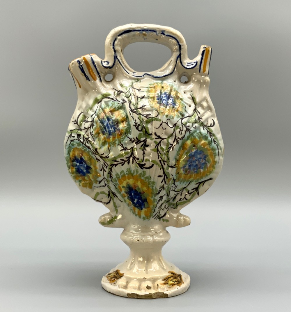An 18th-century Italian polychrome majolica pilgrim flask formed as a scallop shell with an overhead handle, side loops, and hand-painted floral motifs in blue, yellow, and green over a cream glazed surface.