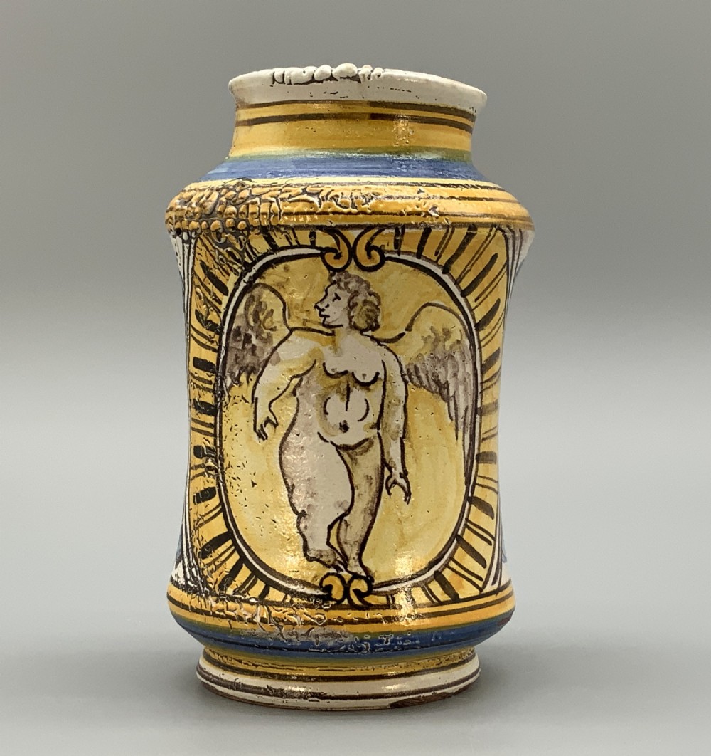 A waisted cylindrical tin-glazed majolica albarello jar decorated in yellow, blue, and dark brown hand-painted pigments featuring a central winged figure motif.