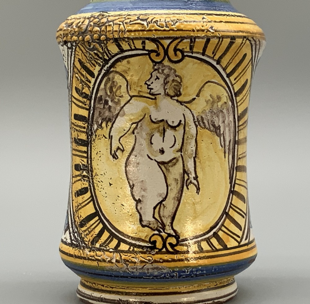 A waisted cylindrical tin-glazed majolica albarello jar decorated in yellow, blue, and dark brown hand-painted pigments featuring a central winged figure motif. View VIII