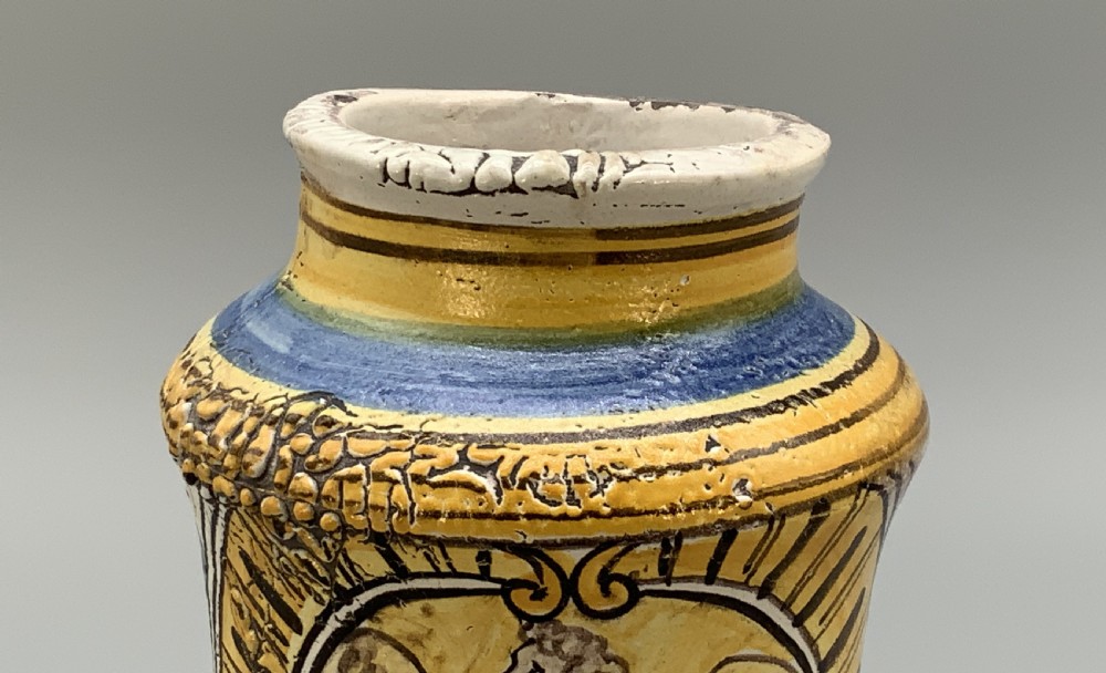 A waisted cylindrical tin-glazed majolica albarello jar decorated in yellow, blue, and dark brown hand-painted pigments featuring a central winged figure motif. View IX