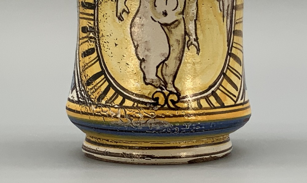 A waisted cylindrical tin-glazed majolica albarello jar decorated in yellow, blue, and dark brown hand-painted pigments featuring a central winged figure motif. View X