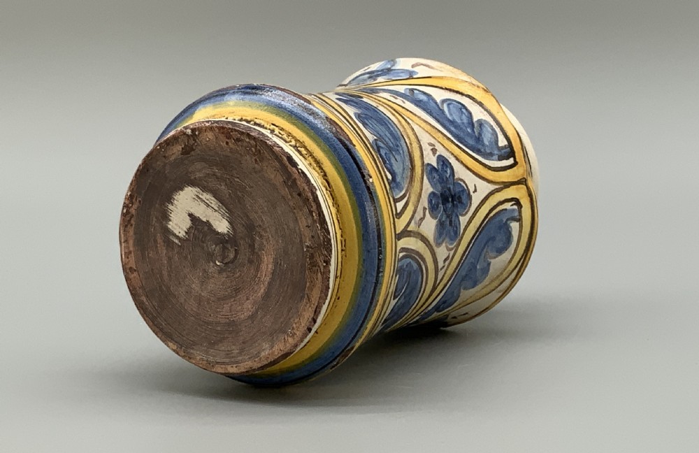 A waisted cylindrical tin-glazed majolica albarello jar decorated in yellow, blue, and dark brown hand-painted pigments featuring a central winged figure motif. View XIII