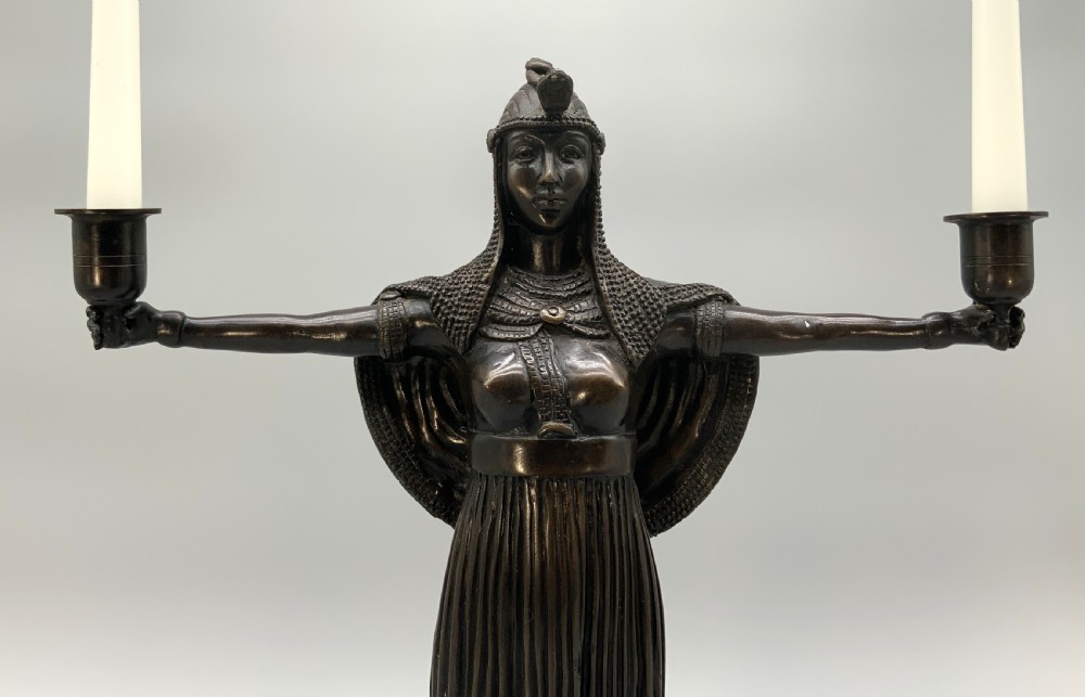 A patinated bronze Egyptian Revival candelabra featuring a female figure with outstretched arms holding two candle sockets, standing on a decorated triangular base. View III