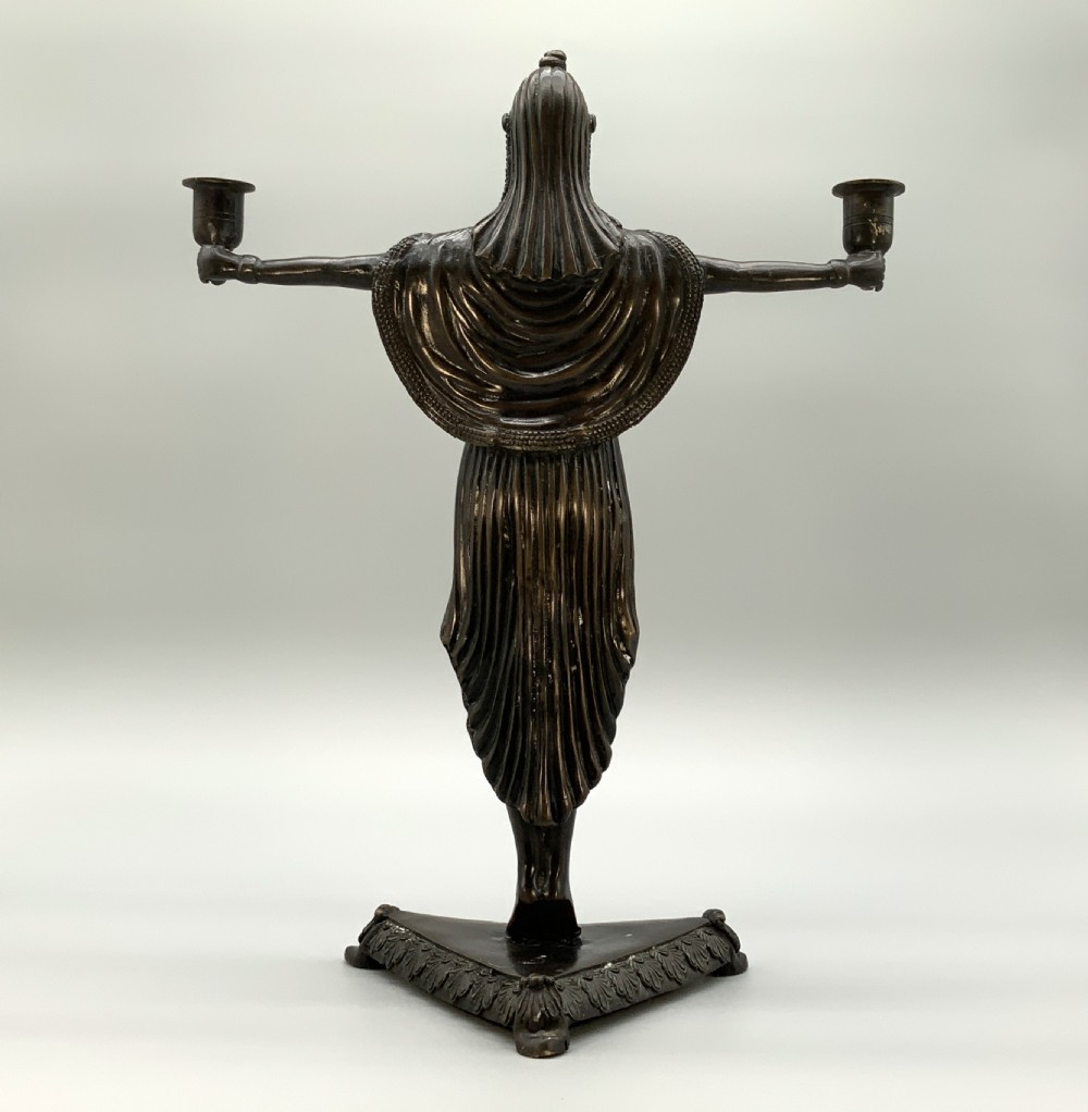 A patinated bronze Egyptian Revival candelabra featuring a female figure with outstretched arms holding two candle sockets, standing on a decorated triangular base. View VI