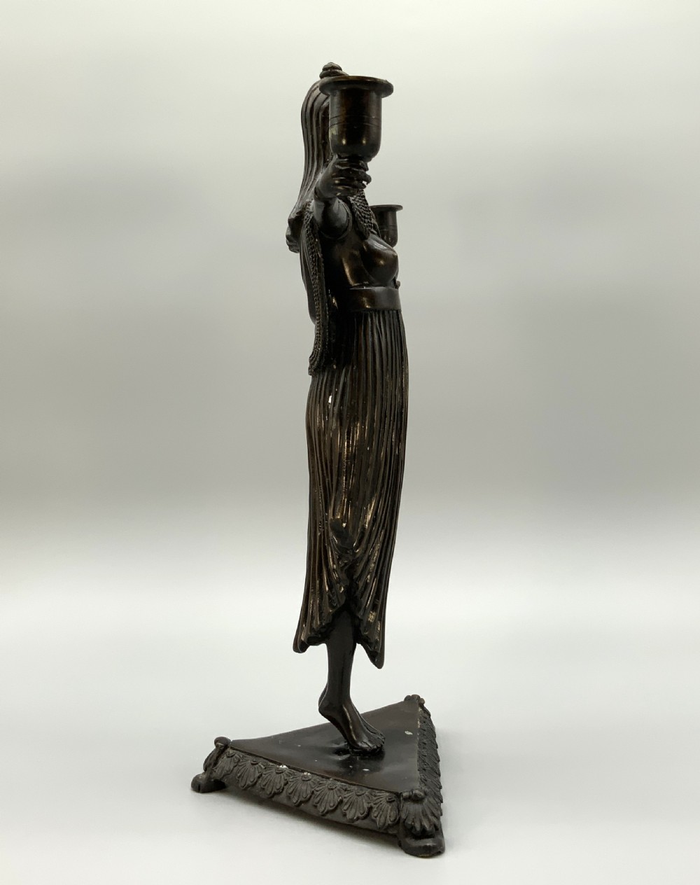 A patinated bronze Egyptian Revival candelabra featuring a female figure with outstretched arms holding two candle sockets, standing on a decorated triangular base. View VII
