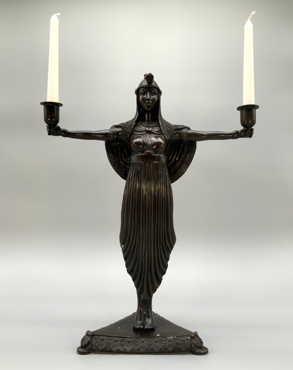 A patinated bronze Egyptian Revival candelabra featuring a female figure with outstretched arms holding two candle sockets, standing on a decorated triangular base. View XIX