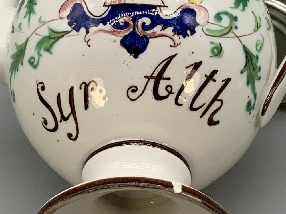An 18th-century Italian tin-glazed earthenware pharmacy wet drug jug with a spouted body, top handle, lid with sculpted finial, painted armorial crest, floral scrolls, and Latin text reading Syr Alth. View XVII