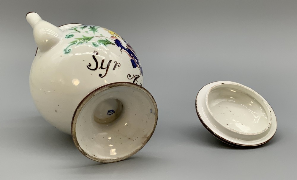 An 18th-century Italian tin-glazed earthenware pharmacy wet drug jug with a spouted body, top handle, lid with sculpted finial, painted armorial crest, floral scrolls, and Latin text reading Syr Alth. View XVIII