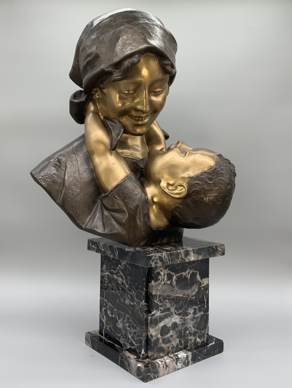 A two-tone bronze bust of a mother wearing a headscarf cradling her infant, mounted on a tall rectangular black veined marble pedestal.
