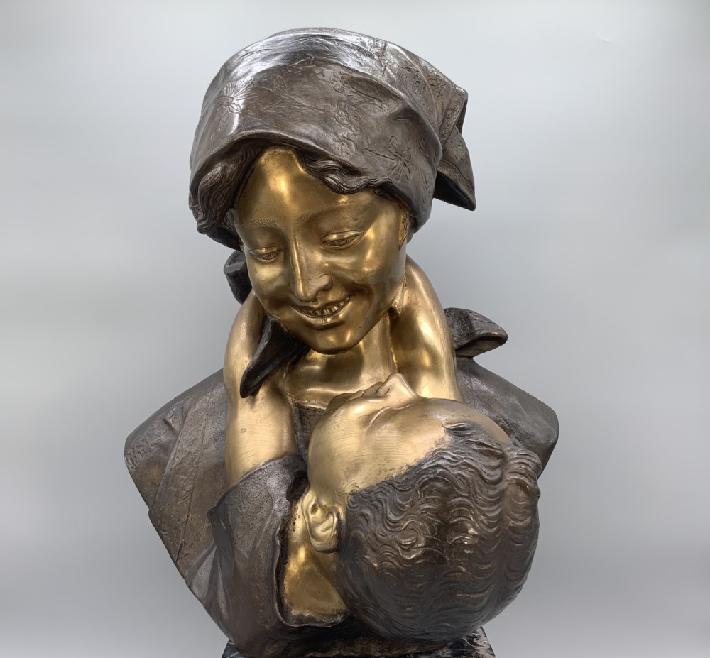 A two-tone bronze bust of a mother wearing a headscarf cradling her infant, mounted on a tall rectangular black veined marble pedestal. View V