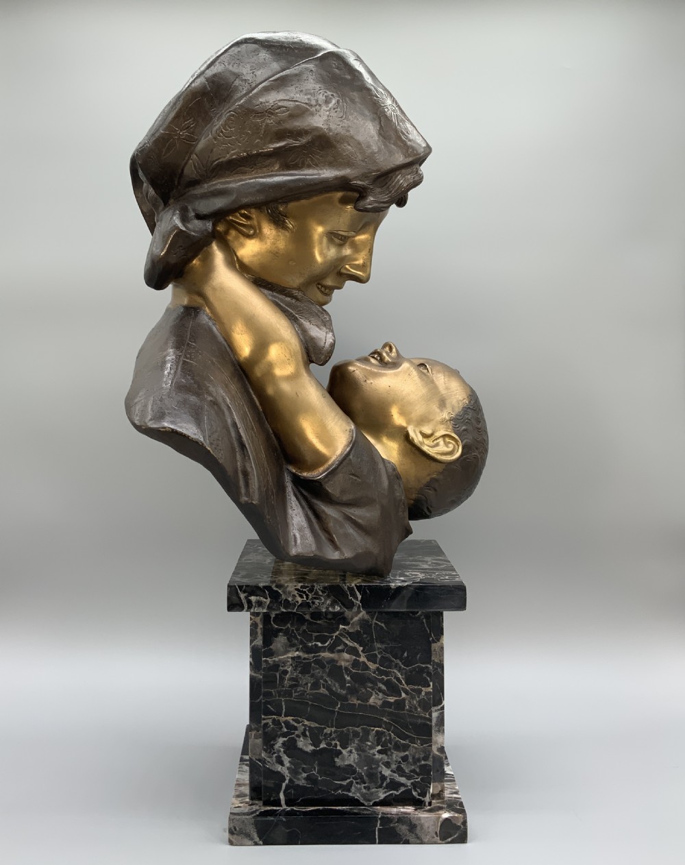 A two-tone bronze bust of a mother wearing a headscarf cradling her infant, mounted on a tall rectangular black veined marble pedestal. View VII