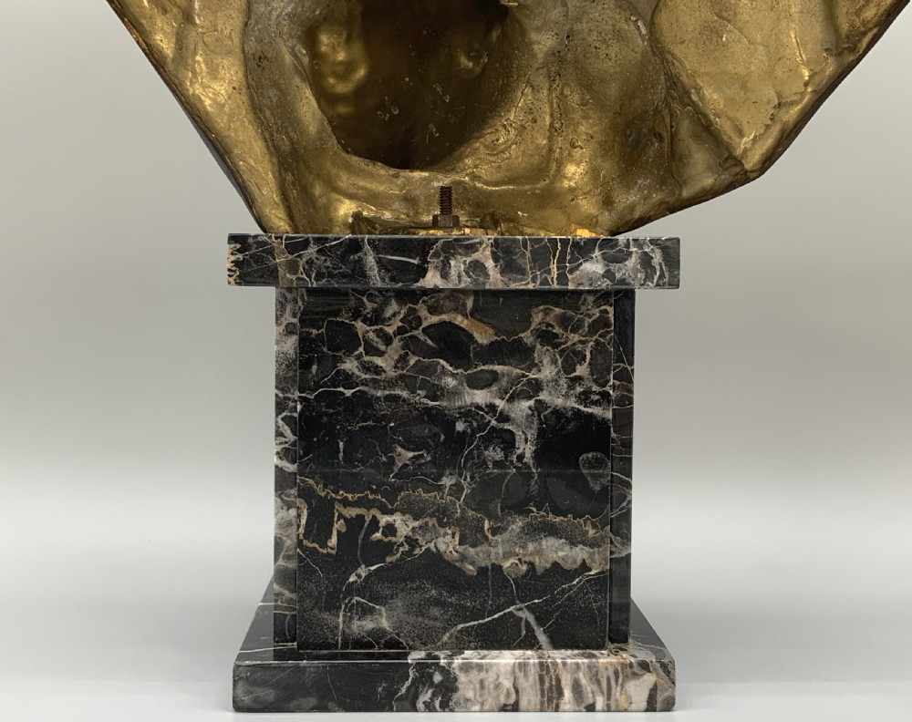 A two-tone bronze bust of a mother wearing a headscarf cradling her infant, mounted on a tall rectangular black veined marble pedestal. View X