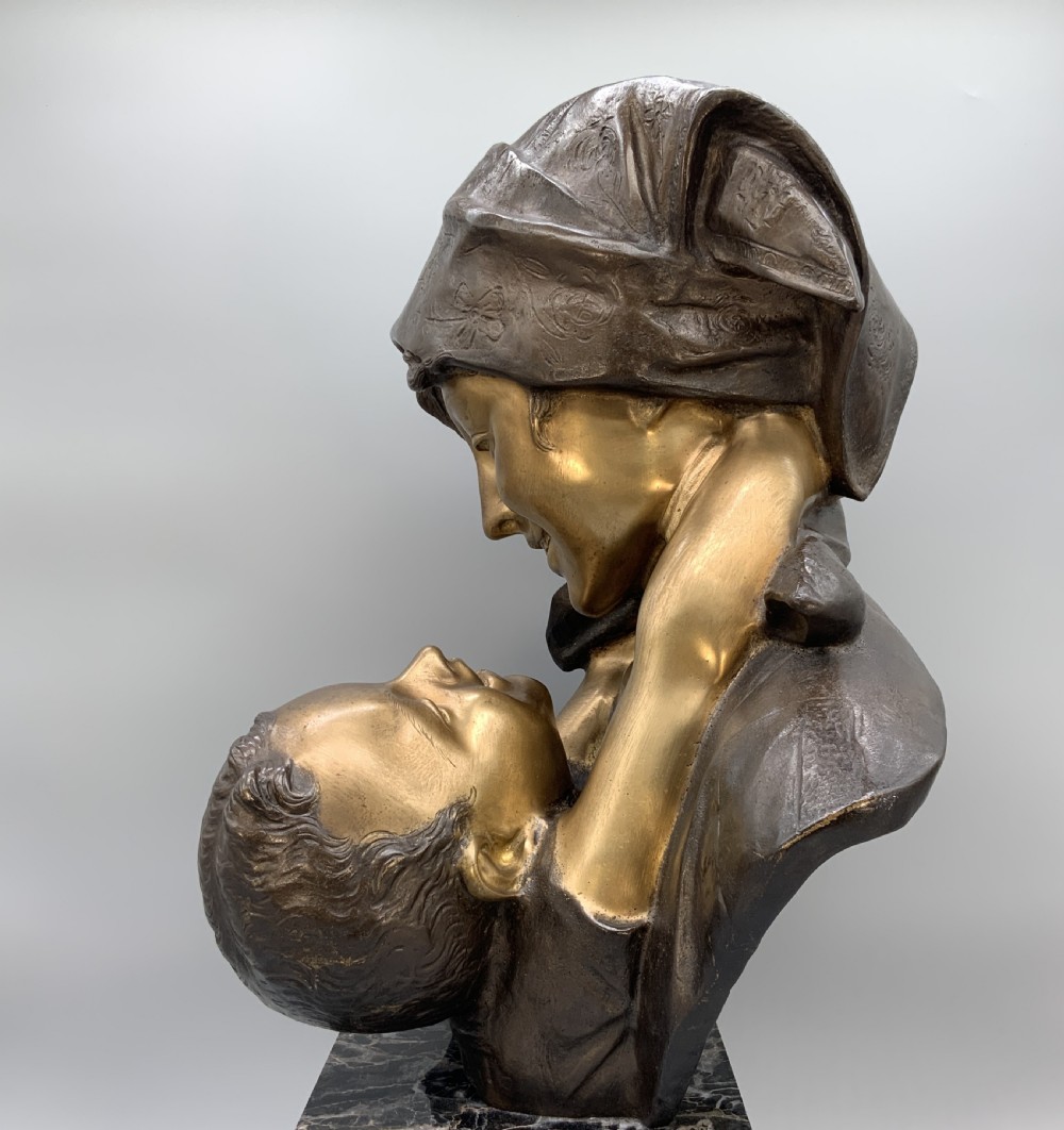A two-tone bronze bust of a mother wearing a headscarf cradling her infant, mounted on a tall rectangular black veined marble pedestal. View XII