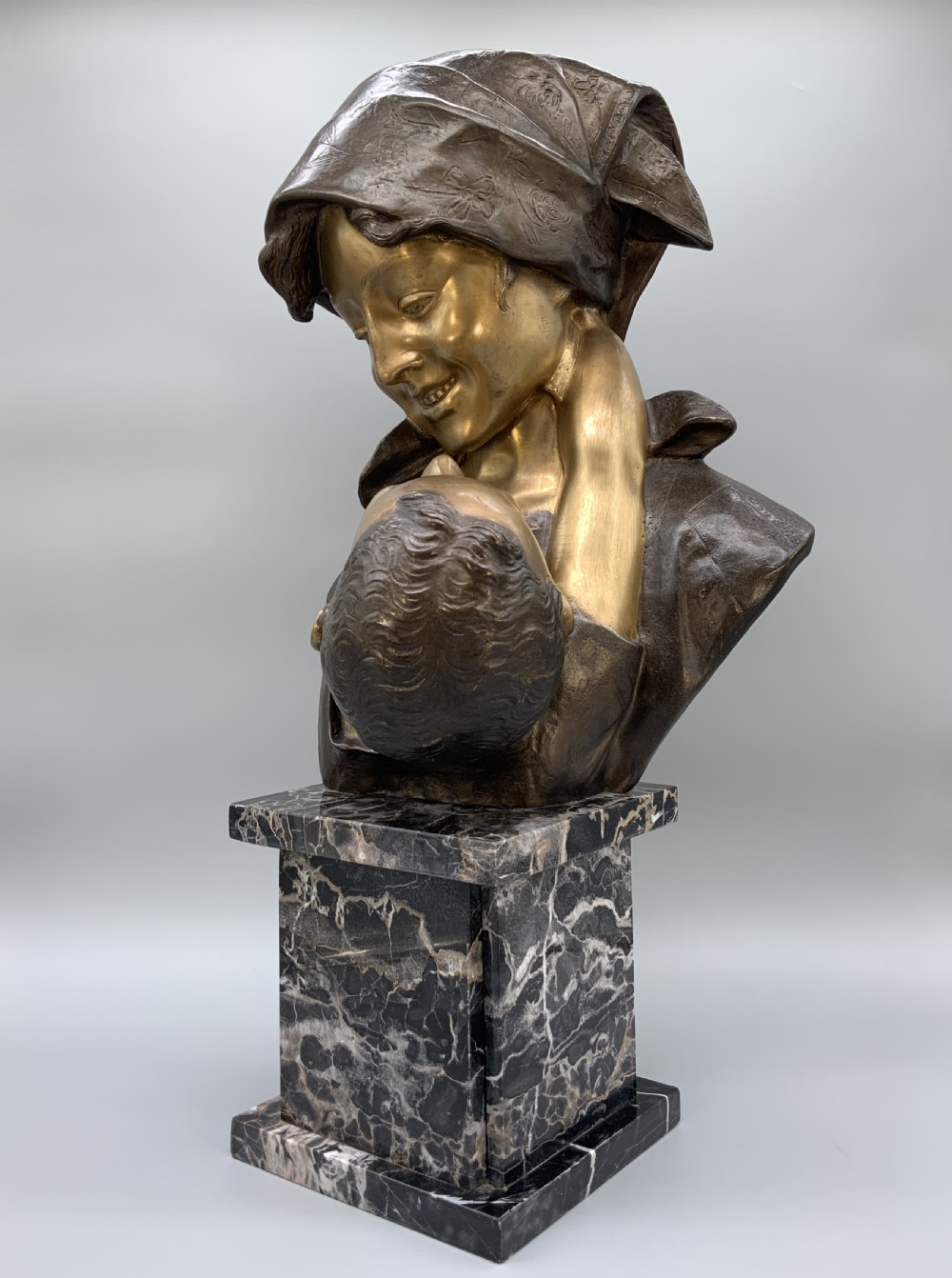 A two-tone bronze bust of a mother wearing a headscarf cradling her infant, mounted on a tall rectangular black veined marble pedestal. View XIII