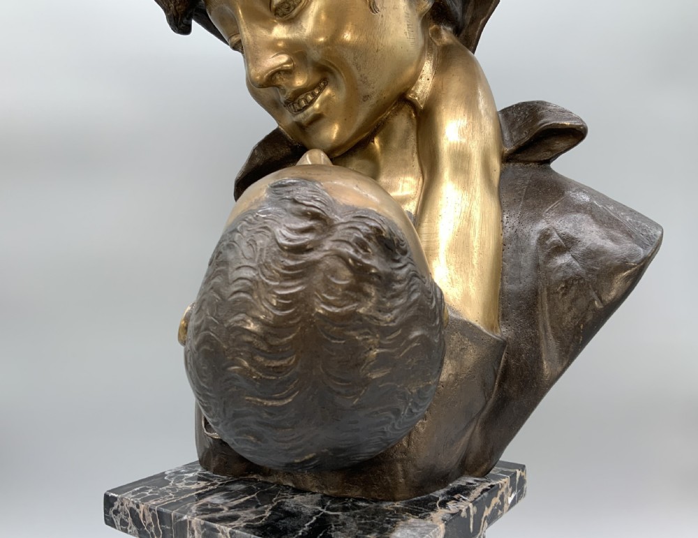 A two-tone bronze bust of a mother wearing a headscarf cradling her infant, mounted on a tall rectangular black veined marble pedestal. View XV