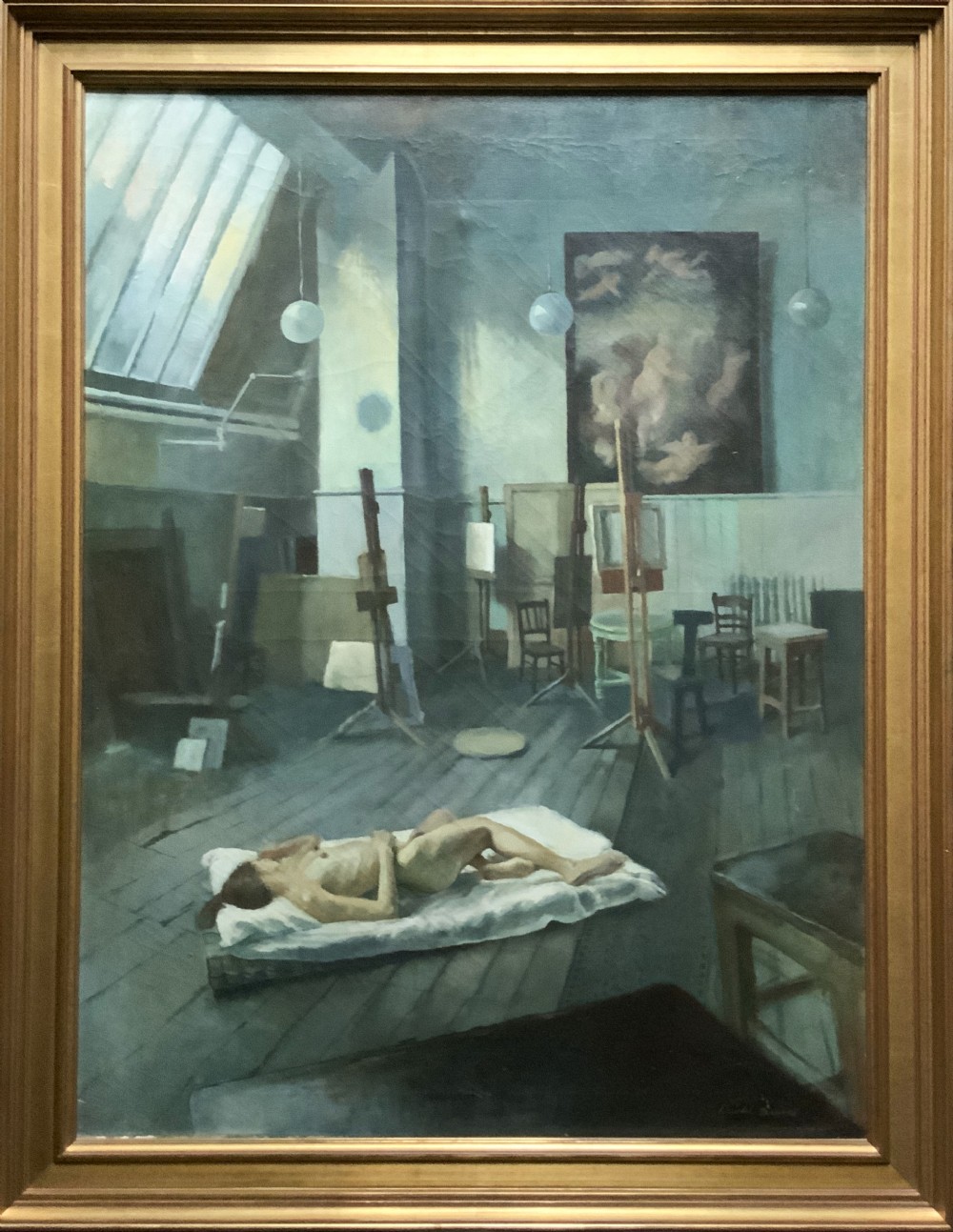 A large framed oil painting depicting an artist studio with a ceiling skylight, wooden easels, and a reclining nude figure on a floor mattress.