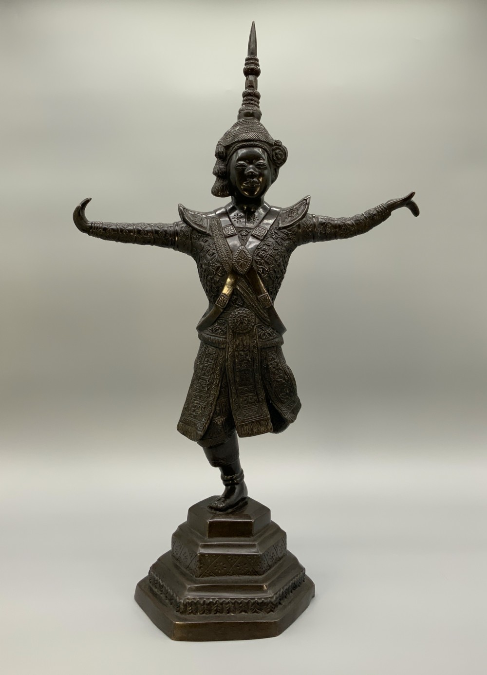 A dark bronze sculpture depicting a classical court dancer in ceremonial attire with a tall spired headdress, balanced on one foot atop an octagonal stepped base.
