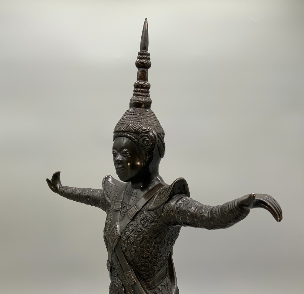 A dark bronze sculpture depicting a classical court dancer in ceremonial attire with a tall spired headdress, balanced on one foot atop an octagonal stepped base. View V