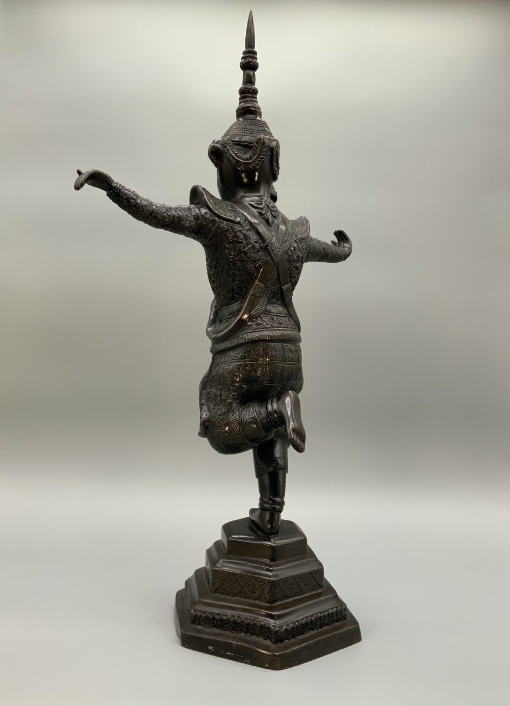 A dark bronze sculpture depicting a classical court dancer in ceremonial attire with a tall spired headdress, balanced on one foot atop an octagonal stepped base. View VI