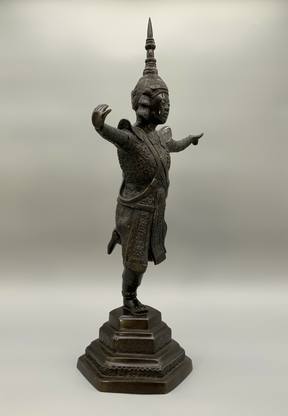 A dark bronze sculpture depicting a classical court dancer in ceremonial attire with a tall spired headdress, balanced on one foot atop an octagonal stepped base. View VIII