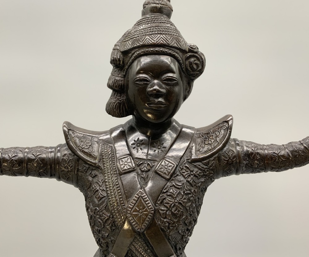 A dark bronze sculpture depicting a classical court dancer in ceremonial attire with a tall spired headdress, balanced on one foot atop an octagonal stepped base. View X