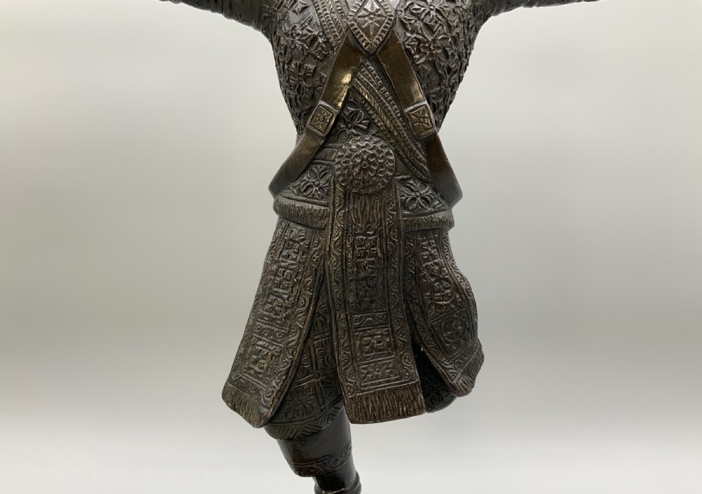 A dark bronze sculpture depicting a classical court dancer in ceremonial attire with a tall spired headdress, balanced on one foot atop an octagonal stepped base. View XI
