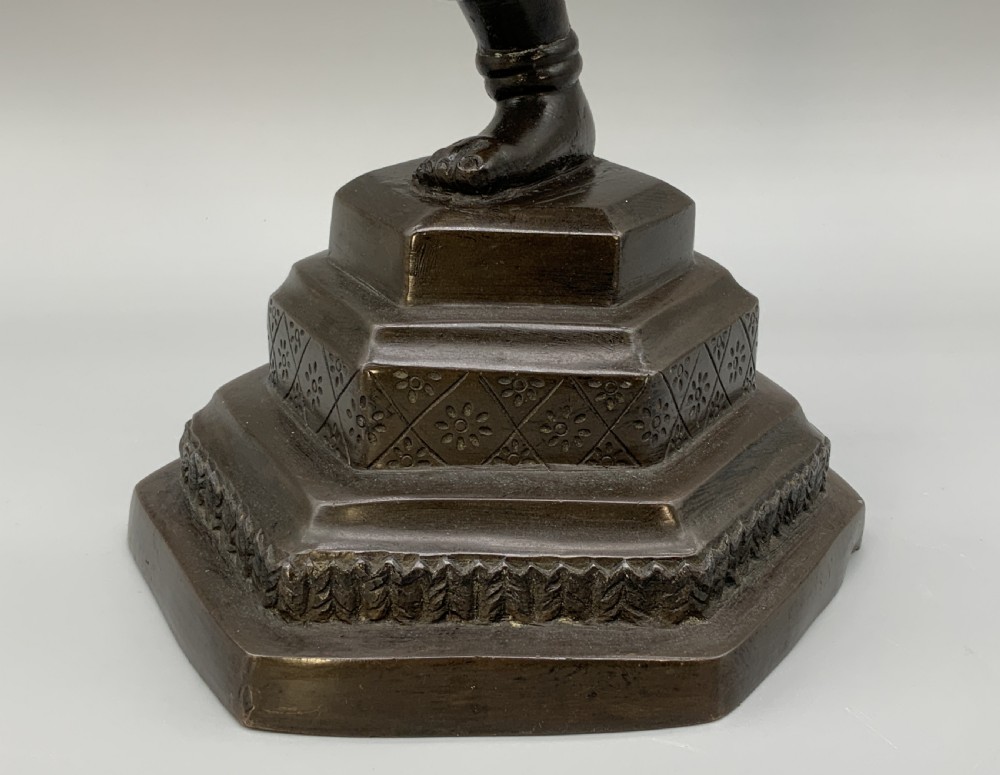 A dark bronze sculpture depicting a classical court dancer in ceremonial attire with a tall spired headdress, balanced on one foot atop an octagonal stepped base. View XII