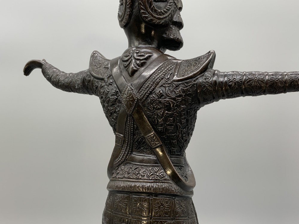 A dark bronze sculpture depicting a classical court dancer in ceremonial attire with a tall spired headdress, balanced on one foot atop an octagonal stepped base. View XVII