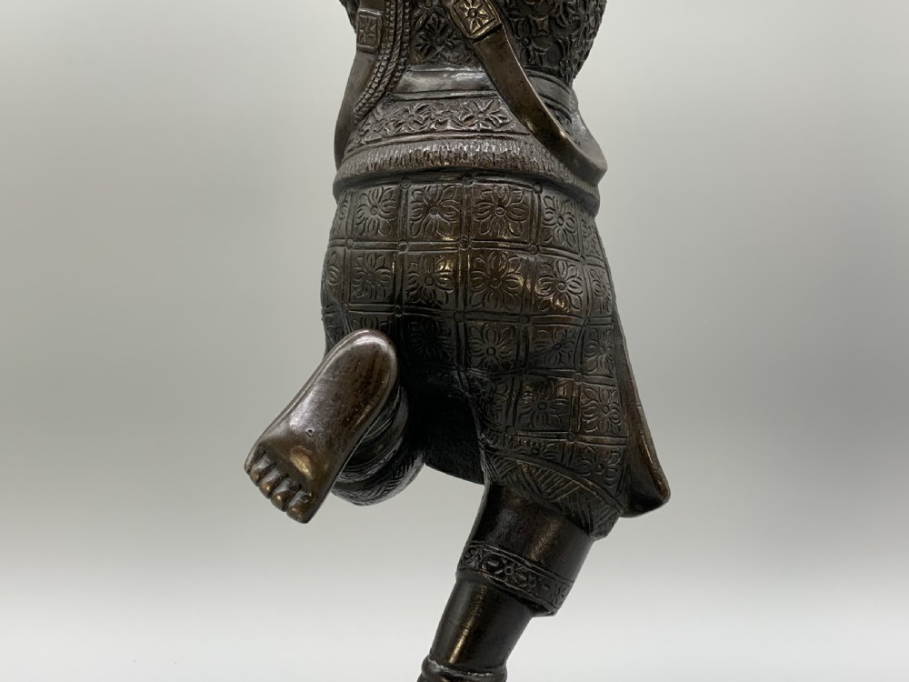 A dark bronze sculpture depicting a classical court dancer in ceremonial attire with a tall spired headdress, balanced on one foot atop an octagonal stepped base. View XVIII