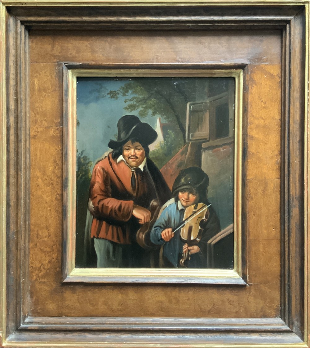 An antique oil painting depicting two musicians, a man and a boy playing a violin, set in a wide wooden frame with a gilded inner moulding.