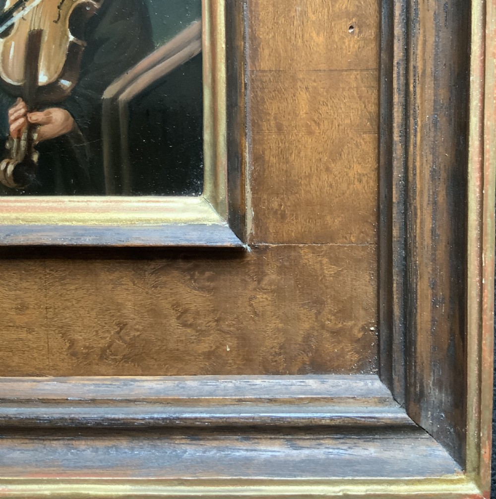 An antique oil painting depicting two musicians, a man and a boy playing a violin, set in a wide wooden frame with a gilded inner moulding. View IX