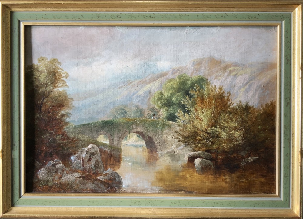 A framed 19th-century oil painting depicting an arched stone bridge crossing a river amidst trees and distant mountains.