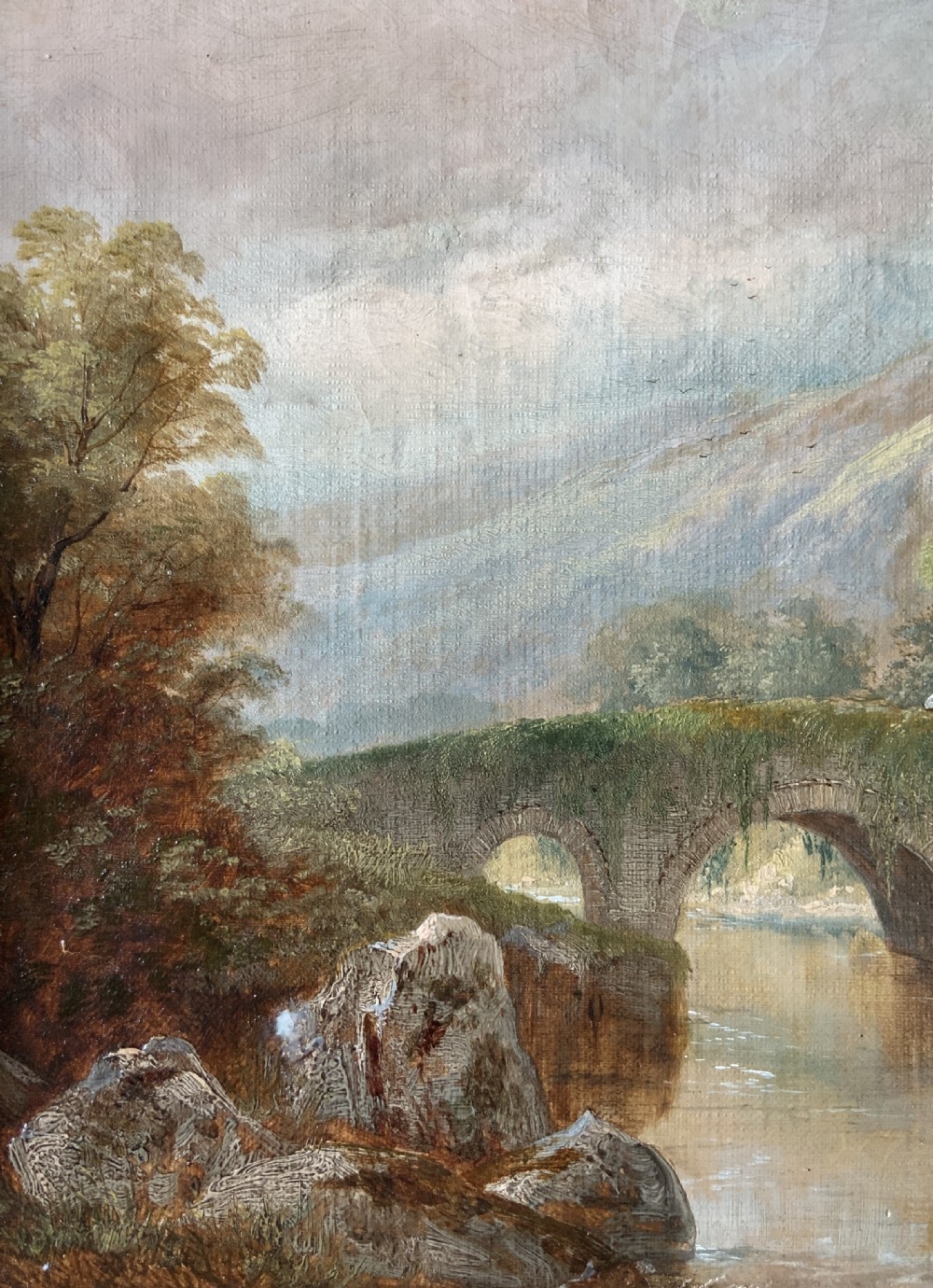 A framed 19th-century oil painting depicting an arched stone bridge crossing a river amidst trees and distant mountains. View V