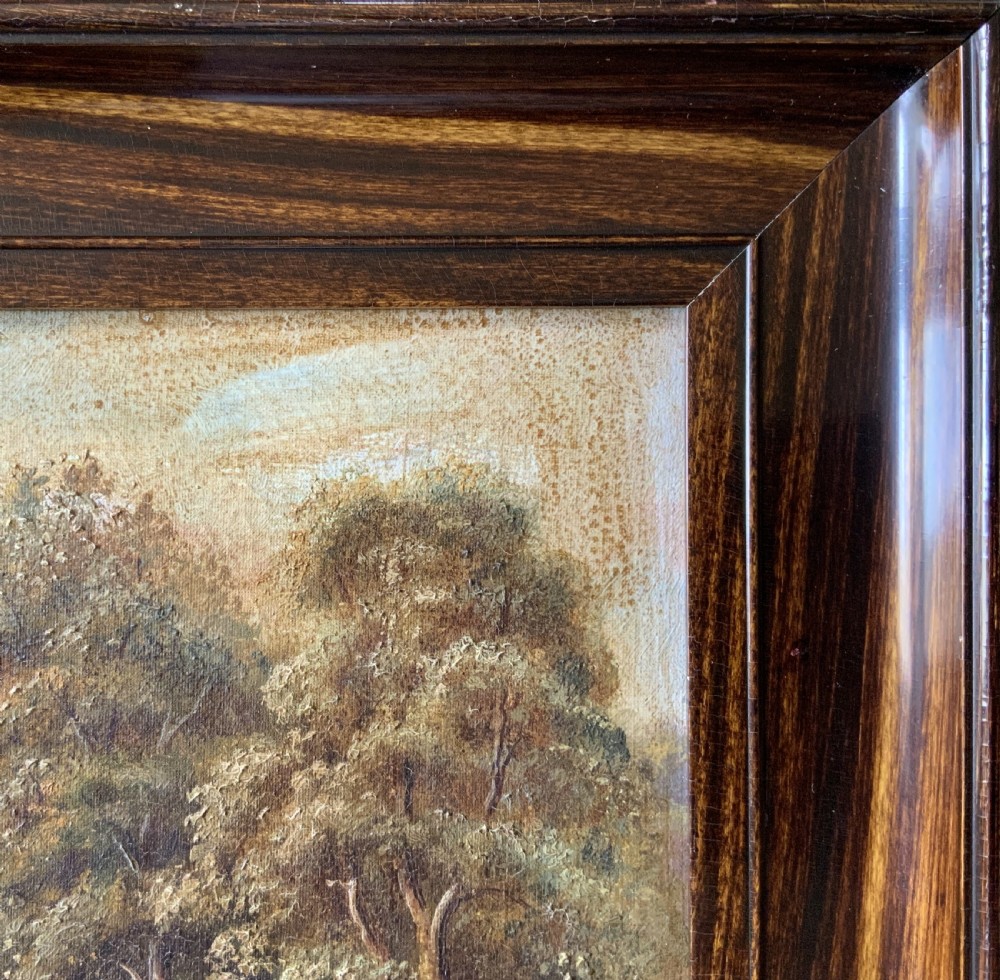 An antique oil painting depicting a tranquil river flowing through a wooded landscape with a stone bridge and cottage in the background, framed in polished dark wood. View XIII