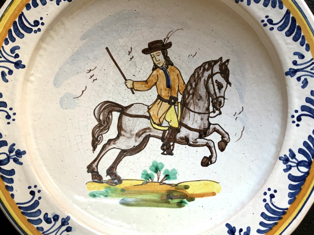 An 18th-century circular Spanish faience charger plate featuring a hand-painted equestrian figure framed by a blue patterned border and a yellow rim. View II