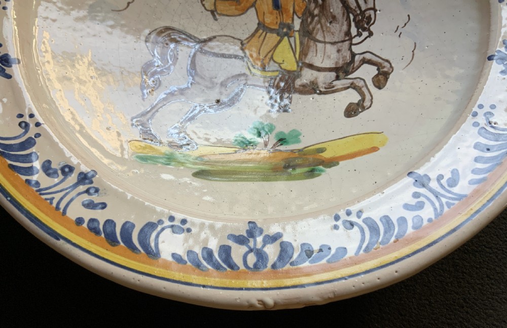An 18th-century circular Spanish faience charger plate featuring a hand-painted equestrian figure framed by a blue patterned border and a yellow rim. View IV