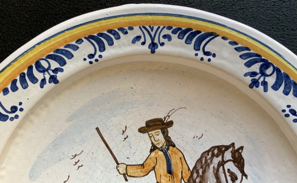 An 18th-century circular Spanish faience charger plate featuring a hand-painted equestrian figure framed by a blue patterned border and a yellow rim. View V