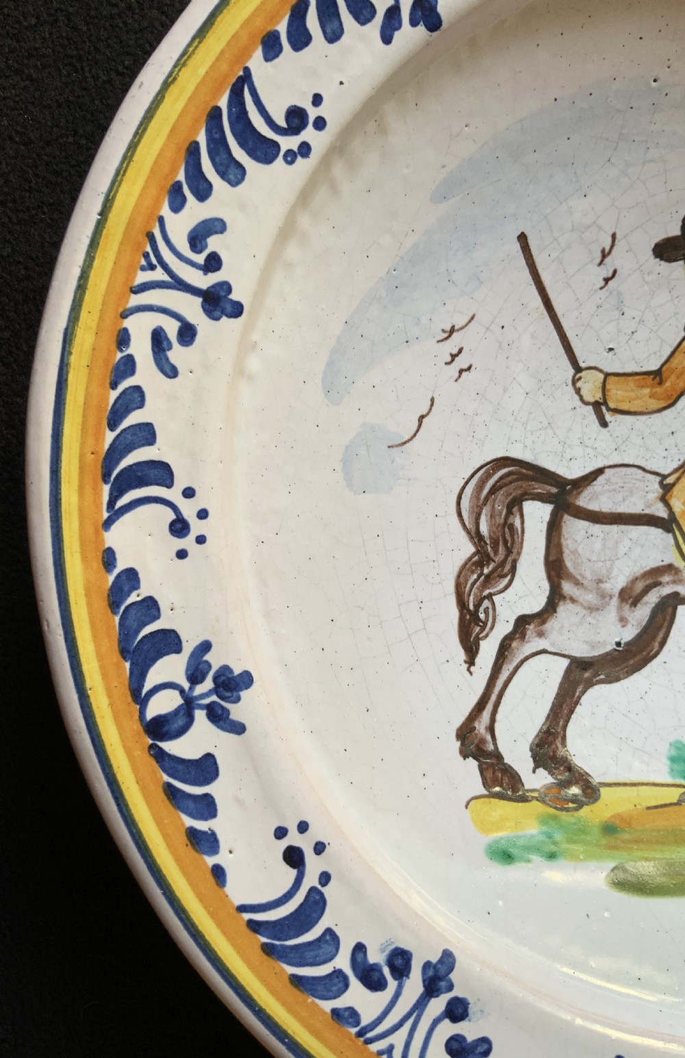 An 18th-century circular Spanish faience charger plate featuring a hand-painted equestrian figure framed by a blue patterned border and a yellow rim. View VI