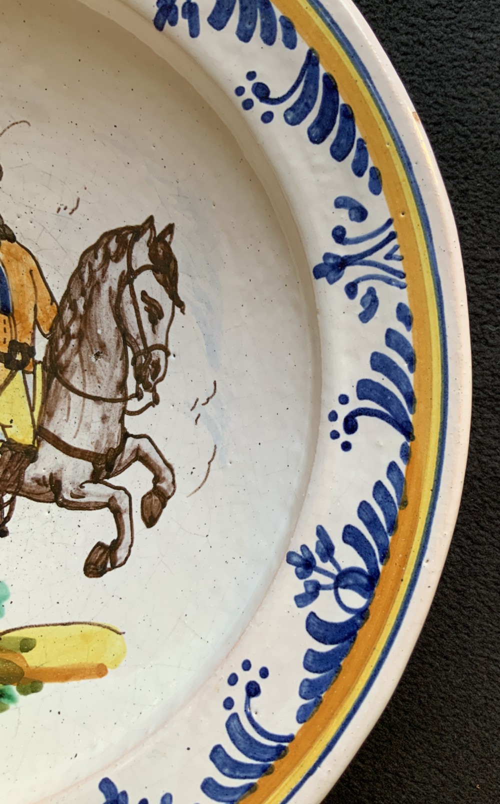 An 18th-century circular Spanish faience charger plate featuring a hand-painted equestrian figure framed by a blue patterned border and a yellow rim. View VII
