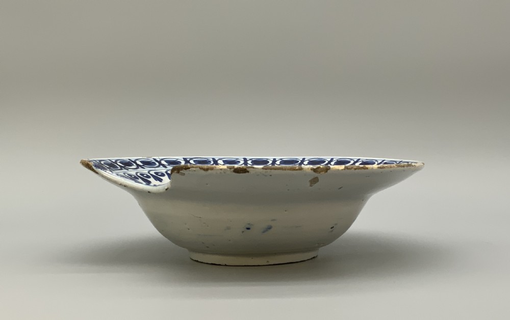 An 18th-century blue and white Dutch Delftware barbers bowl featuring hand-painted floral motifs and a semi-circular rim notch. View X