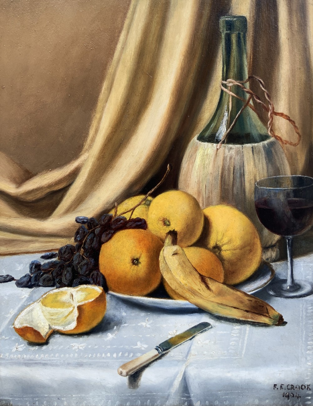 An Edwardian still life oil painting depicting citrus fruit, bananas, grapes, and a glass of wine on a tablecloth, set inside a heavy gold gilded frame. View II