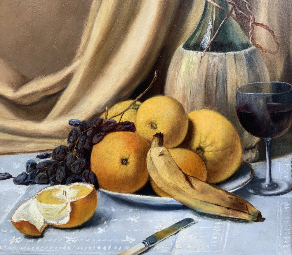 An Edwardian still life oil painting depicting citrus fruit, bananas, grapes, and a glass of wine on a tablecloth, set inside a heavy gold gilded frame. View V