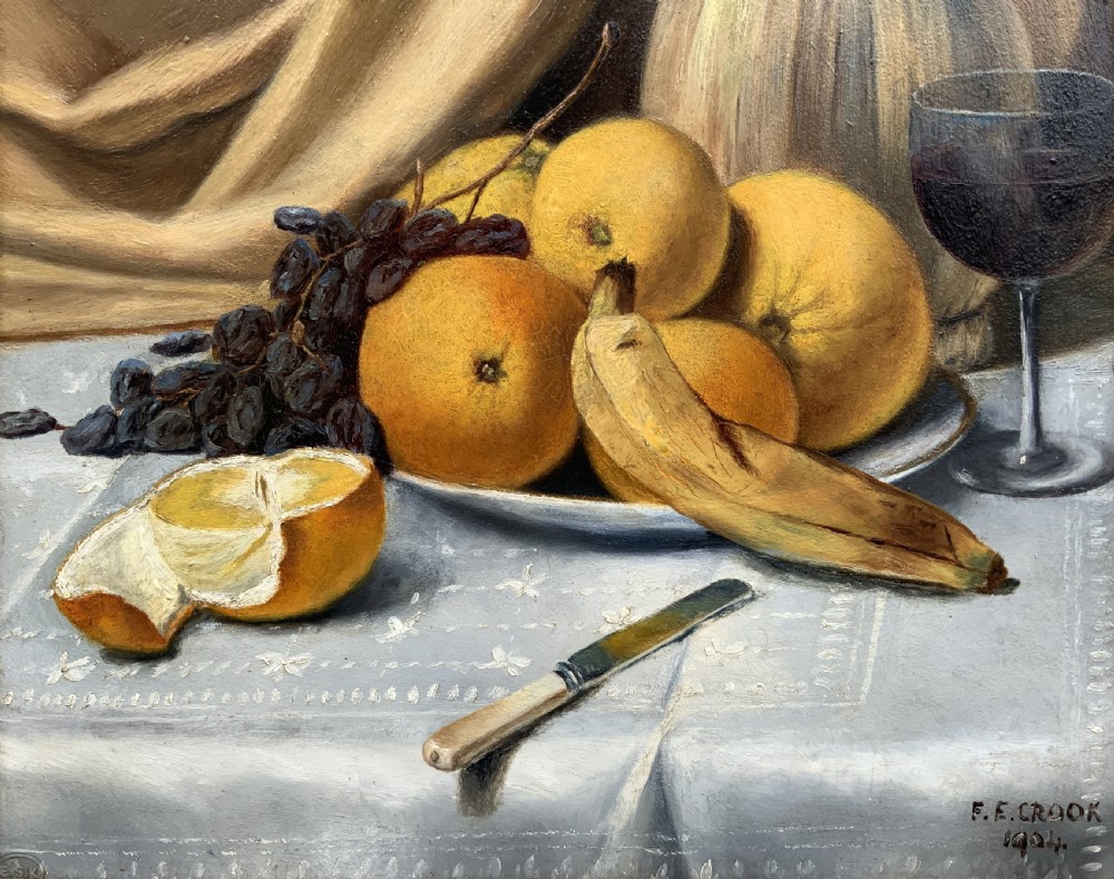 An Edwardian still life oil painting depicting citrus fruit, bananas, grapes, and a glass of wine on a tablecloth, set inside a heavy gold gilded frame. View VI