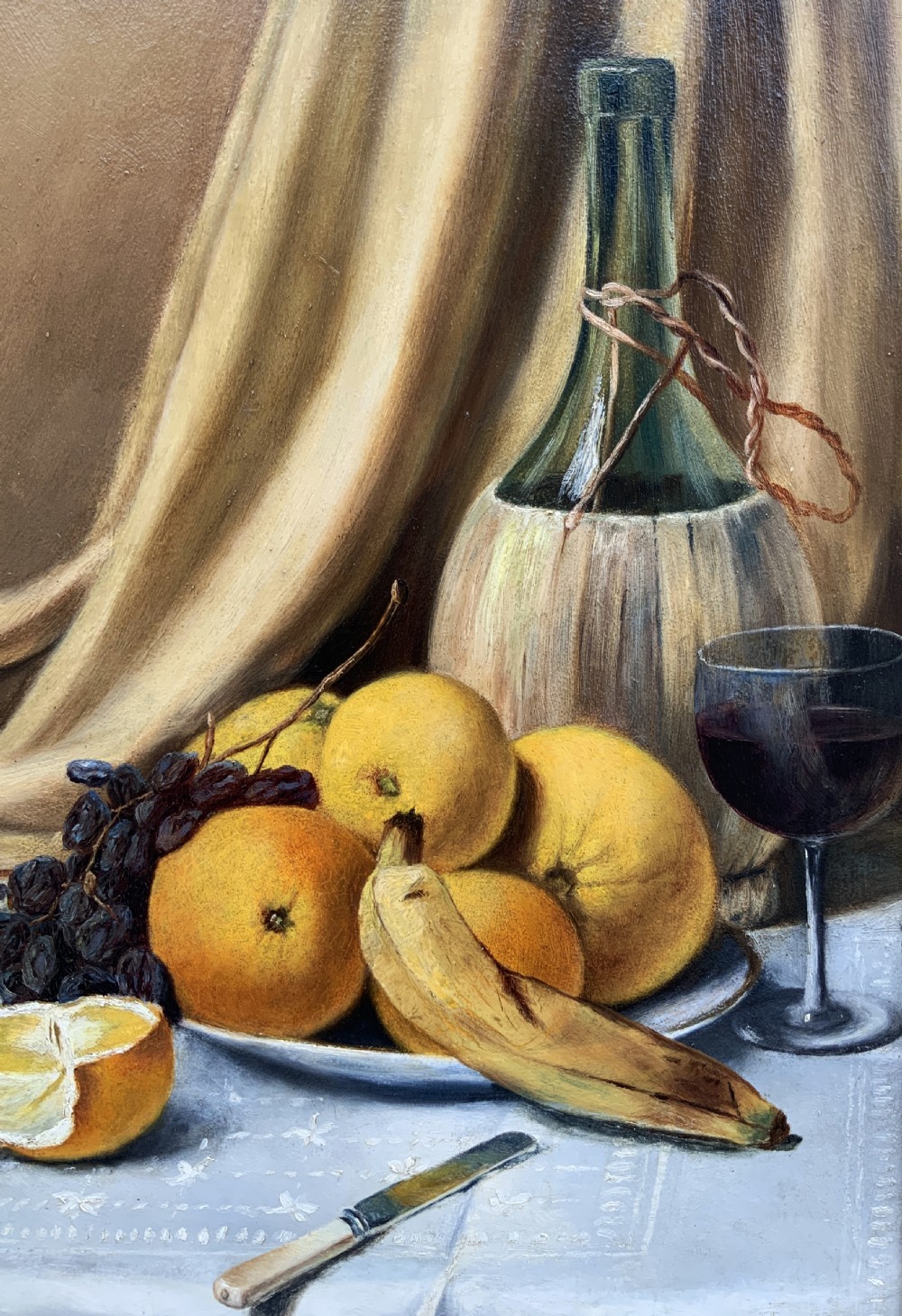 An Edwardian still life oil painting depicting citrus fruit, bananas, grapes, and a glass of wine on a tablecloth, set inside a heavy gold gilded frame. View VII