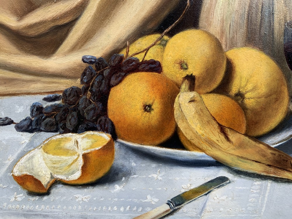 An Edwardian still life oil painting depicting citrus fruit, bananas, grapes, and a glass of wine on a tablecloth, set inside a heavy gold gilded frame. View VIII